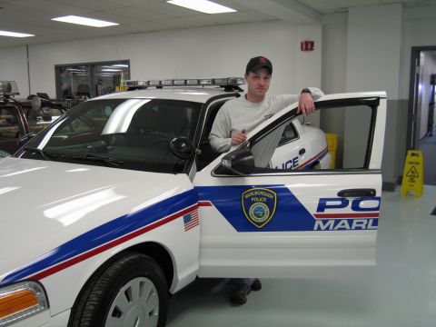1991 Ford LTD Crown Victoria - man in a cap stands at the open driver's door of a white 'MARLBOROUGH POLICE' cruiser with a roof light bar in a showroom.