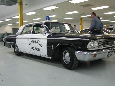 1991 Ford LTD Crown Victoria - black-and-white 1960s Ford 'HAMILTON POLICE' sedan with a single blue dome light and chrome trim, parked in a showroom.