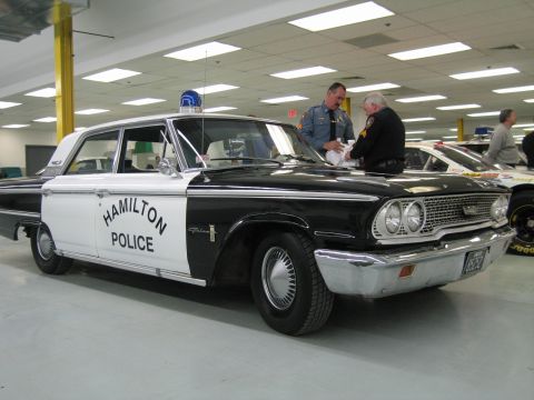 1991 Ford LTD Crown Victoria - Two uniformed men stand at the hood of a black-and-white 1960s 'HAMILTON POLICE' Ford sedan with a blue dome light indoors.