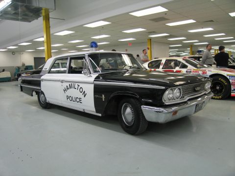 1991 Ford LTD Crown Victoria - Front three-quarter view of a black-and-white 1960s Ford 'HAMILTON POLICE' sedan with blue dome light in a showroom.