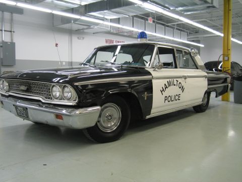 1991 Ford LTD Crown Victoria - restored black-and-white vintage Ford sedan marked Hamilton Police, with a single blue roof beacon, parked indoors on a polished floor.