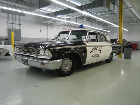 1991 Ford LTD Crown Victoria - Front three-quarter view of a black-and-white vintage Ford sedan lettered Hamilton Police with a blue dome light, displayed in a shop.