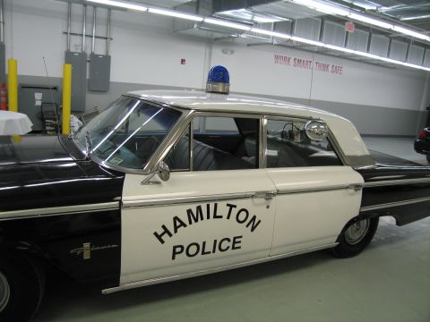 1991 Ford LTD Crown Victoria - Close side view of a black-and-white sedan's door painted HAMILTON POLICE, with a chrome side mirror and blue roof beacon.