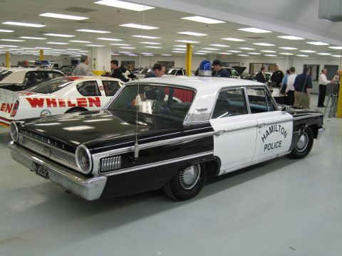 1991 Ford LTD Crown Victoria - Rear three-quarter view of a black-and-white Hamilton Police Ford sedan, parked beside a red Whelen-liveried race car in a shop.