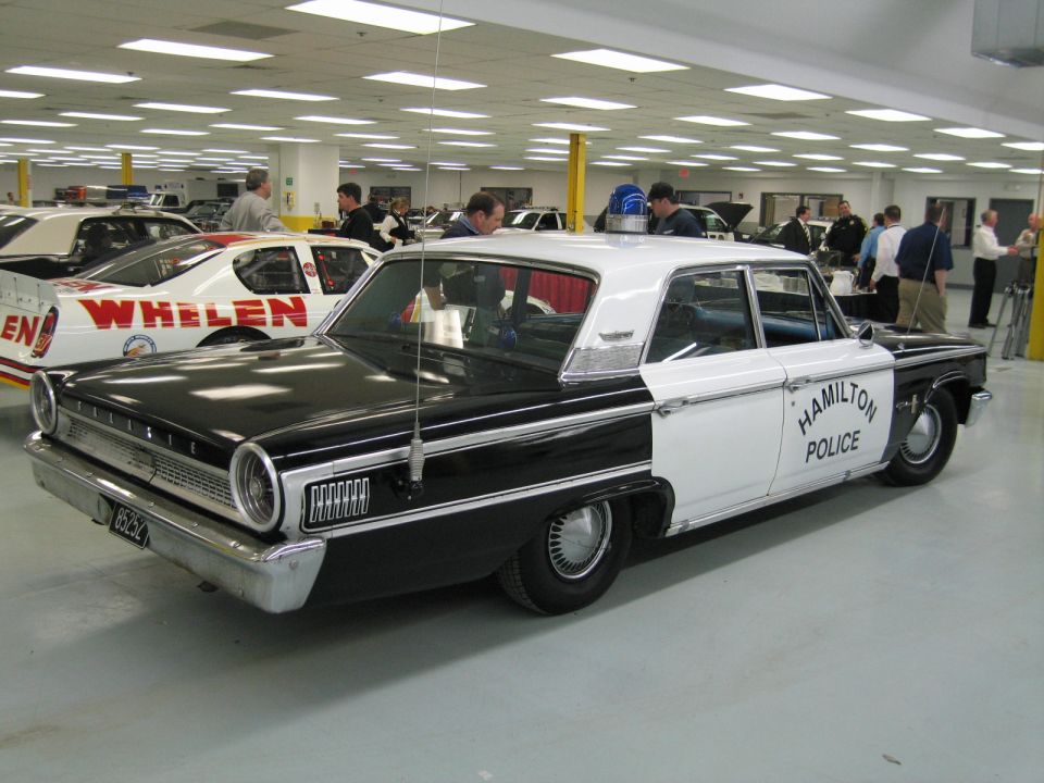 Rear view of black and white Ford police sedan near a red Whelen race car indoors