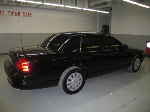 1991 Ford LTD Crown Victoria - Rear three-quarter view of a glossy black Ford sedan with one illuminated taillight, parked against a wall reading THINK SAFE.