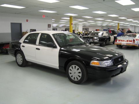 1991 Ford LTD Crown Victoria - Front three-quarter view of a black-and-white Ford sedan with handwritten window notes, parked near a red Whelen race car.