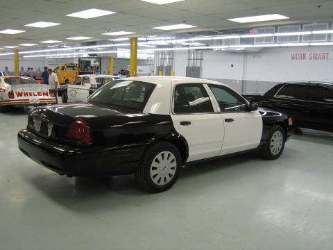 1991 Ford LTD Crown Victoria - Rear three-quarter view of a black-and-white Ford sedan parked in a large shop near a Whelen race car and a WORK SMART sign.