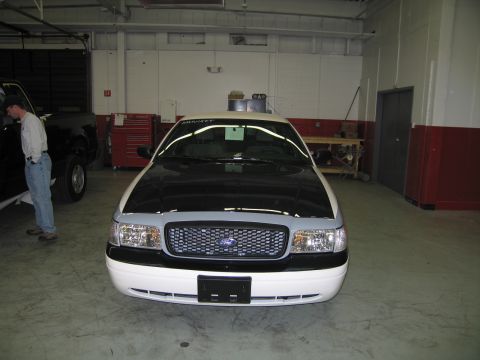 1991 Ford LTD Crown Victoria - Head-on front view of a white Ford sedan with a chrome mesh grille and roof light bar, with a man standing to the left in a garage.