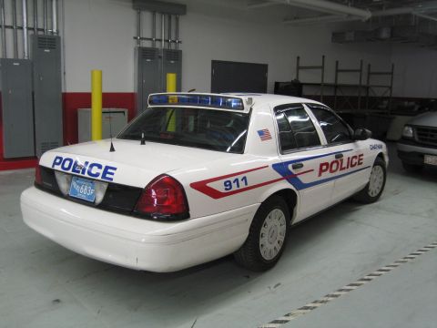 1991 Ford LTD Crown Victoria - Rear three-quarter view of a white police car marked POLICE, 911 and an American-flag decal, parked in a garage near lockers.