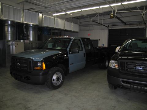 1991 Ford LTD Crown Victoria - dark flatbed Ford dually pickup sits beside a darker Ford pickup inside a metal-walled garage, both showing Ford grille badges.