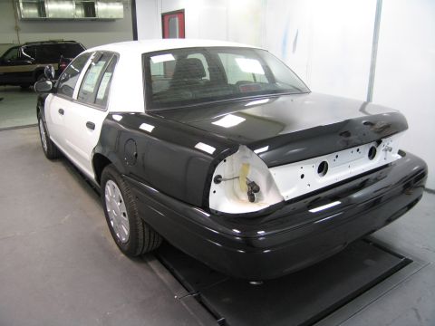 1991 Ford LTD Crown Victoria - rear of a black-and-white sedan sits on a lift in a paint booth, its trunk lid primed and rear bumper removed exposing mounting holes.
