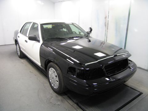 1991 Ford LTD Crown Victoria - black-and-white sedan sits on a lift in a spray booth with its front bumper removed and front end coated in dark primer.