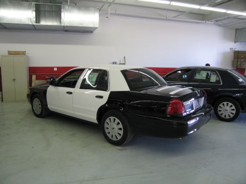 1991 Ford LTD Crown Victoria - glossy black-and-white police sedan is parked at an angle in a shop next to a dark sedan, with a red stripe along the wall behind.