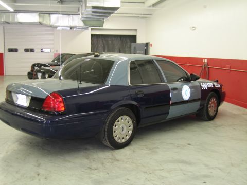 1991 Ford LTD Crown Victoria - two-tone blue sedan with a Massachusetts seal and "500 STATE POLICE" door lettering sits in a fleet garage with a red wall stripe.