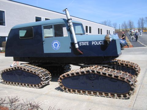 1991 Ford LTD Crown Victoria - blue "STATE POLICE" Tucker Sno-Cat with an exhaust stack and four tracks sits on pavement outside a long modern building.