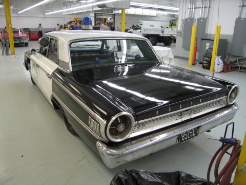 1991 Ford LTD Crown Victoria - rear of a black-and-white Ford Galaxie shows its "GALAXIE" trim, round taillights and chrome bumper inside a busy workshop.