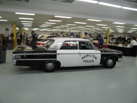 1991 Ford LTD Crown Victoria - black-and-white "Hamilton Police" Ford sedan sits in profile in a warehouse-style showroom as people work in the background.