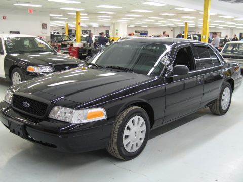1991 Ford LTD Crown Victoria - black Ford Crown Victoria sedan with a pillar spotlight sits at a front angle in a large workshop alongside other police-style sedans.