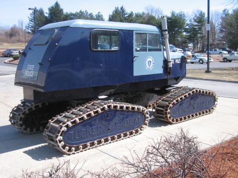 1991 Ford LTD Crown Victoria - blue tracked snow vehicle with a State Police seal sits on a curb, its Tucker Sno-Cat tracks visible against a wooded backdrop.