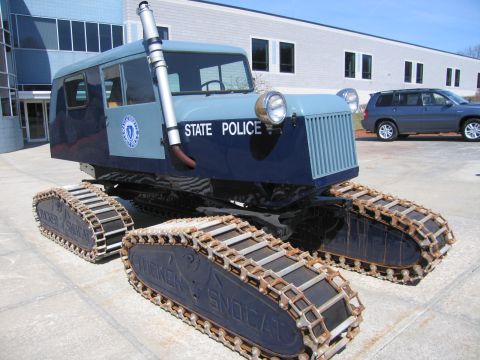 1991 Ford LTD Crown Victoria - blue tracked snow vehicle lettered STATE POLICE with a large headlight and exhaust stack sits on pavement with a blue SUV behind it.