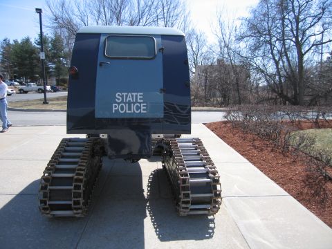 1991 Ford LTD Crown Victoria - rear of a blue tracked snow vehicle stenciled STATE POLICE, with bare trees and mulch beds in the background.