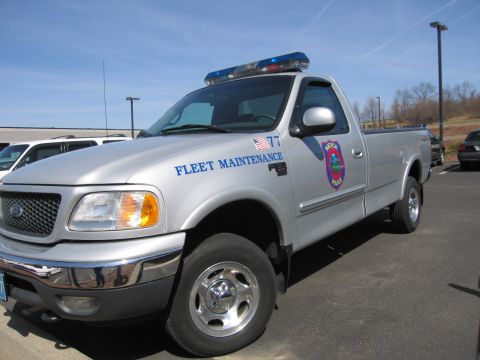 1991 Ford LTD Crown Victoria - silver Ford F-150 pickup marked FLEET MAINTENANCE with a roof light bar and police emblem is parked in a lot.