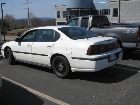 1991 Ford LTD Crown Victoria - white Chevrolet Impala sedan with black steel wheels is parked beside a dark pickup outside a modern building.