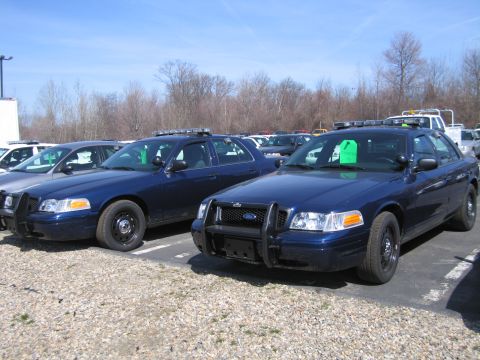 1991 Ford LTD Crown Victoria - Two dark blue Ford Crown Victoria police cruisers, one with a front push bar, parked among rows of cars before bare trees.