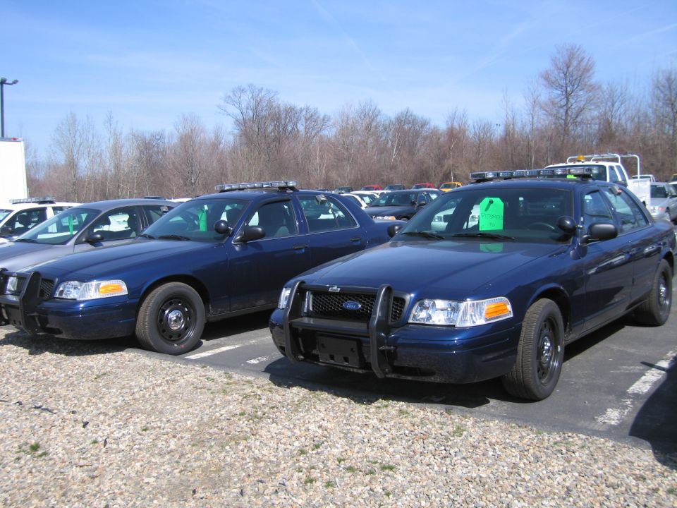 Two dark blue Ford Crown Victoria police cruisers parked side by side on gravel