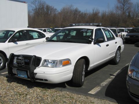 1991 Ford LTD Crown Victoria - white Ford Crown Victoria police interceptor with a front push bar and roof light bar sits among other vehicles on gravel.