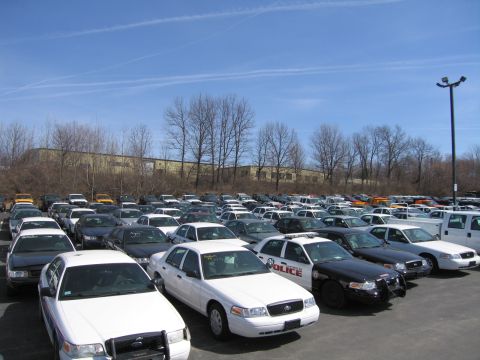 1991 Ford LTD Crown Victoria - Rows of parked police sedans fill a lot, including a black-and-white cruiser marked POLICE, with a warehouse behind bare trees.