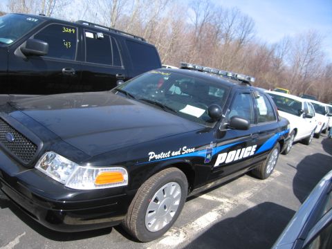 1991 Ford LTD Crown Victoria - black Ford police interceptor sedan with blue striping and 'Protect and Serve POLICE' lettering parked among other vehicles.