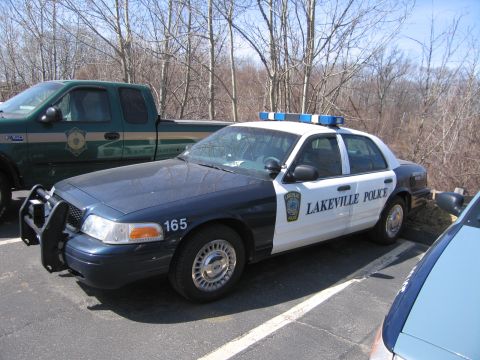 1991 Ford LTD Crown Victoria - dark-blue-and-white Lakeville Police Ford sedan numbered 165 with push bar and light bar, parked near a green pickup.
