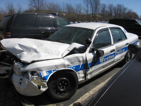 1991 Ford LTD Crown Victoria - white-and-blue Weston Police Ford sedan with severe front-end and side crash damage, missing wheel, parked in a salvage lot.