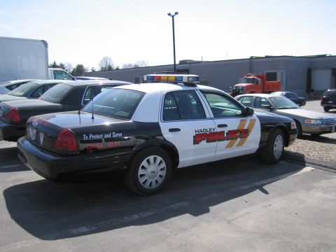 1991 Ford LTD Crown Victoria - Rear three-quarter view of a black-and-white Hadley Police Ford sedan with amber light bar and 'To Protect and Serve' lettering.