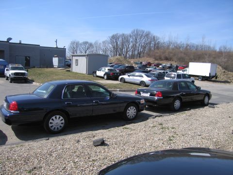 1991 Ford LTD Crown Victoria - Two dark full-size sedans in the foreground with rows of fleet vehicles and a box truck beyond, in a gravel-and-pavement yard.