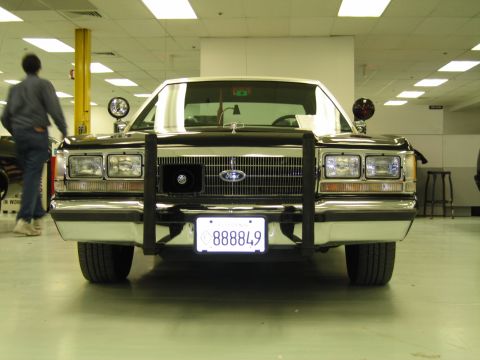 1991 Ford LTD Crown Victoria - Head-on front view of a dark vintage Ford sedan with chrome push bar, dual spotlights and California plate 888849, displayed indoors.