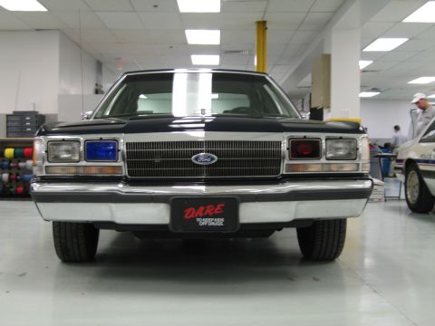 1991 Ford LTD Crown Victoria - Head-on front view of a black vintage Ford sedan with chrome bumper and a red 'D.A.R.E.' front plate, displayed indoors.