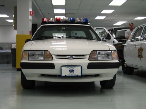 1991 Ford LTD Crown Victoria - white fox-body Ford Mustang police interceptor with a roof-mounted light bar faces the camera indoors, a Sheriff cruiser visible behind it.