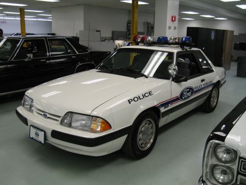 1991 Ford LTD Crown Victoria - white fox-body Ford Mustang marked Police with a roof light bar and Ford Police Vehicles graphics sits on display indoors.