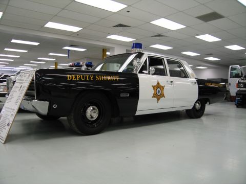 1991 Ford LTD Crown Victoria - black-and-white Deputy Sheriff police sedan with a blue roof light and spotlights sits on the showroom floor beside an info placard.