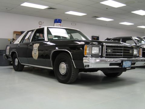 1991 Ford LTD Crown Victoria - black-and-white Ford LTD police sedan with a blue roof light and a police door emblem sits on display in an indoor collection.