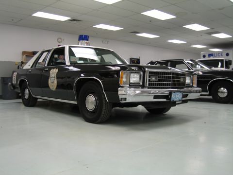 1991 Ford LTD Crown Victoria - black-and-white Ford LTD police cruiser with a gold door emblem and blue roof light sits among other patrol cars indoors.