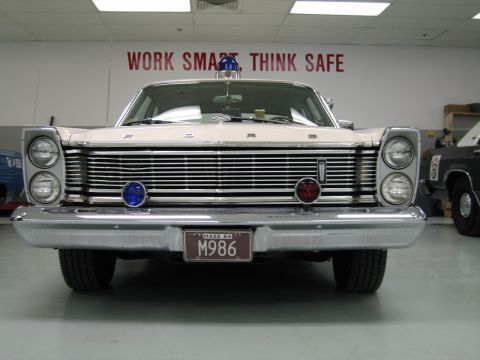 1991 Ford LTD Crown Victoria - front of a white 1960s Ford Galaxie police car with blue dome lights sits below a red Work Smart Think Safe wall sign.