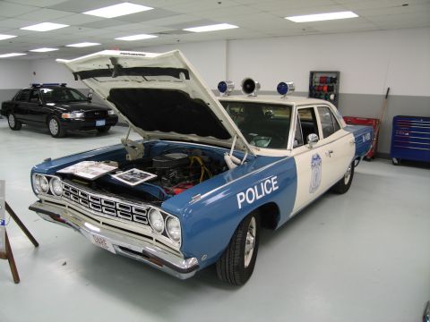 1991 Ford LTD Crown Victoria - blue-and-white Plymouth police car sits with its hood open, engine visible, beside a red rolling tool chest in a garage-like hall.