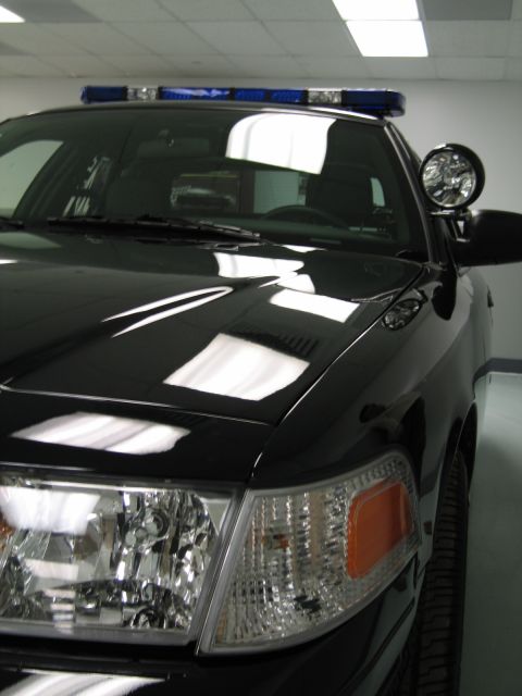 1991 Ford LTD Crown Victoria - close-up of a black Ford Crown Victoria police car shows its fender, headlight, spotlight and blue roof light bar indoors.