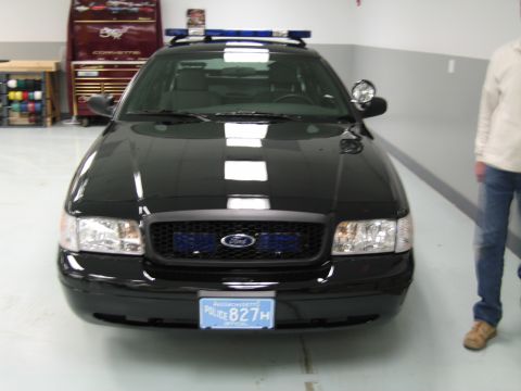1991 Ford LTD Crown Victoria - black Ford Crown Victoria police car faces the camera with blue grille lights and a roof light bar, a person standing at right.