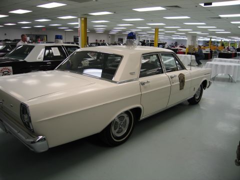 1991 Ford LTD Crown Victoria - rear quarter of a white 1960s Ford Galaxie police sedan with a blue dome light and gold door emblem is shown indoors.