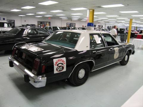 1991 Ford LTD Crown Victoria - black-over-white 1980s full-size police sedan with a blue light bar and a round Emergency 911 decal on the rear fender.